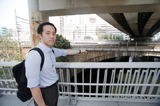 上をよこぎっているのが、1960年代（昭和35～44年）につくられた首都高速道路（しゅとこうそくどうろ）で、太田さんが指をさしているのが大正7年に作られた鉄道アーチ橋（きょう）。そして太田さんが立っている場所が2018年（平成30年）にできた「竜閑さくら橋」。ここでは、大正、昭和、平成と3つの時代の橋を一度に見ることができる。こうした歴史も調べて設計に生かすようにしている。